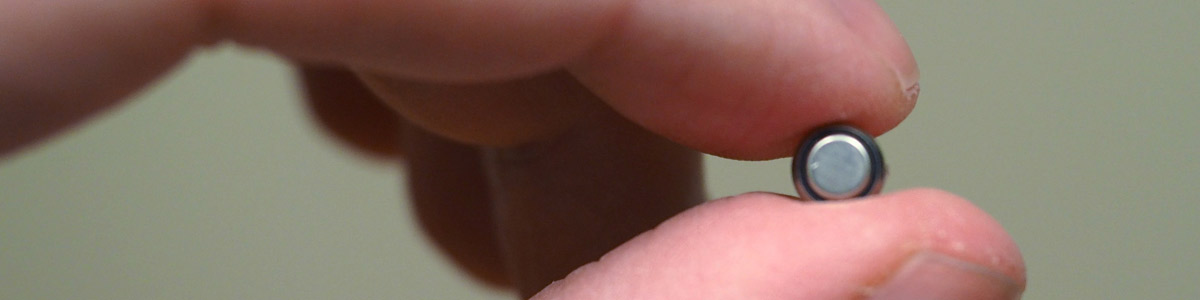 Picture of a very small lithium button battery being pinched between a thumb and index finger to show how little it is. 